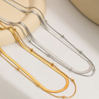 Dos collares, uno dorado y el otro plateado que tiene doble cadena con bolitas encima de una mesa color hueso