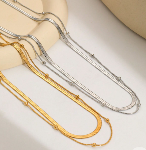 Dos collares, uno dorado y el otro plateado que tiene doble cadena con bolitas encima de una mesa color hueso