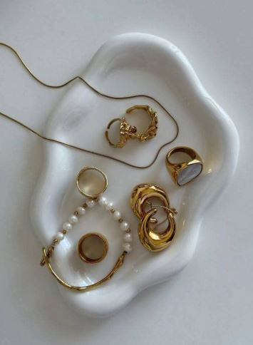Joyas minimalistas de oro y una pulsera con perlas blancas, están encima de un plato en forma abstracta de color blanco