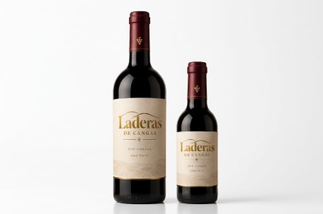 Fotografía de producto de dos botellas de vino tinto 'Laderas de Cangas' DOP Cangas, una de 750 ml y otra más pequeña de 250 ml, presentadas juntas sobre una superficie de madera y una peana beige con fondo claro, mostrando su etiqueta beige y cápsula granate.