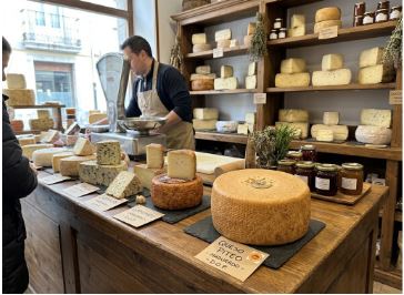 Interior de una tienda de quesos Afuega´l Pitu, donde podemos ver en la parte delantera todos los tipos y piezas de quesos de la marca. En la parte trasera se aprecia la nueva crema de queso con manzana asturiana. Y entre ambos el dependiente de la empresa se encuentra atendiendo a un cliente, pesando el queso en una pesa bastante grande.
